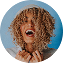 Woman with curly hair laughing with eyes closed against a blue circle background.