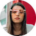 Woman with long brown hair wearing orange-tinted sunglasses against a bright red background.