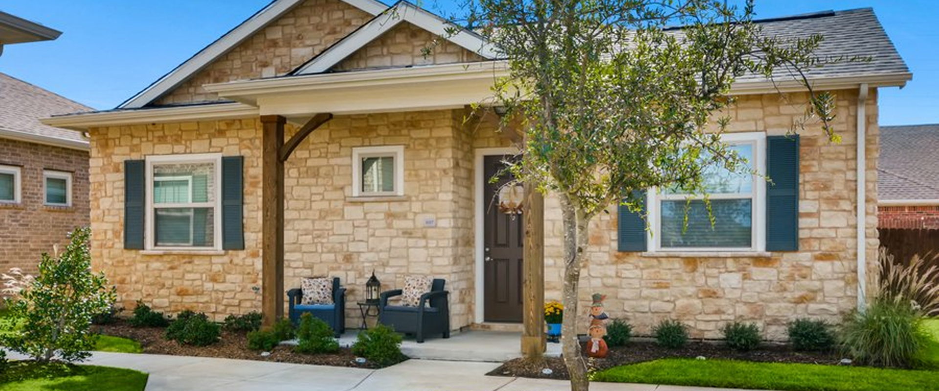 Stone house with dark shutters, front porch with two chairs, small tree, and green lawn.