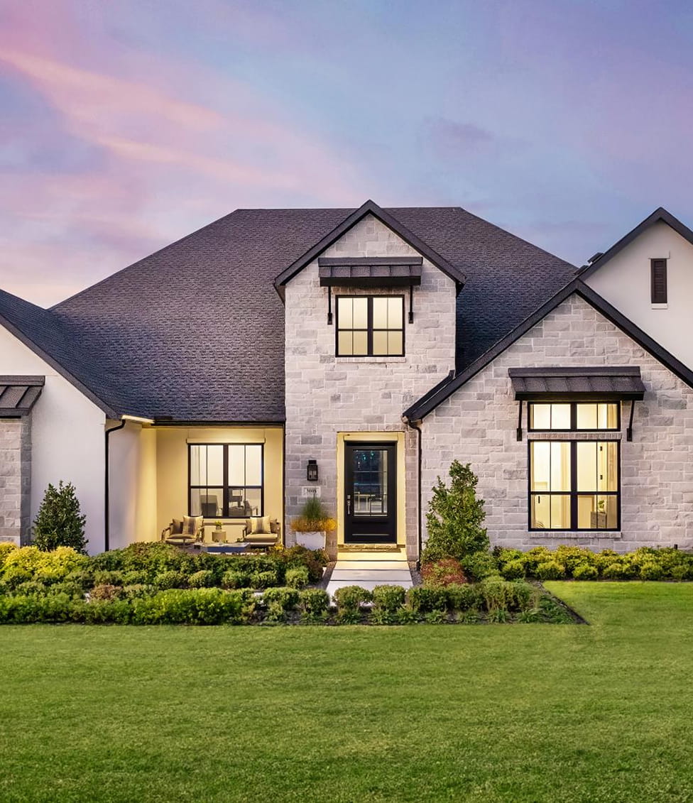 Modern stone house with black roof, large windows, front porch, and manicured green lawn at dusk.