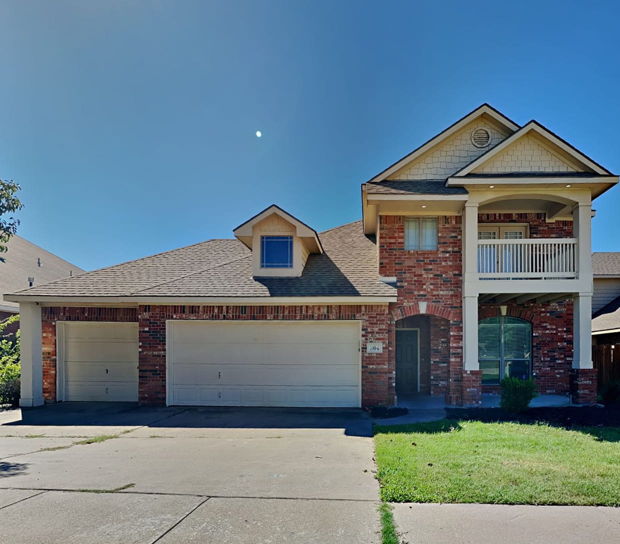 Two-story brick house with three-car garage, front porch balcony, and a green lawn under a clear blue sky.