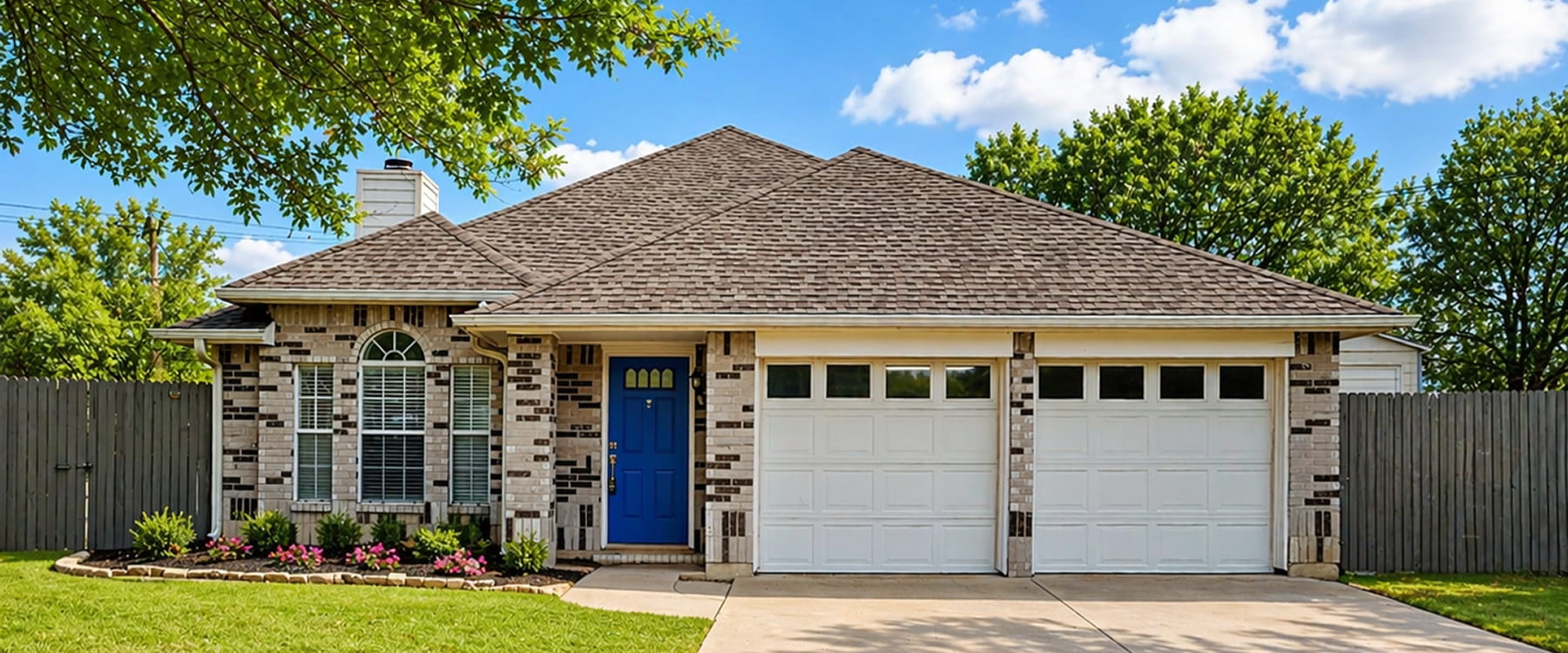 Single-story brick house with gray roof, blue front door, two-car garage, and front yard flowers.
