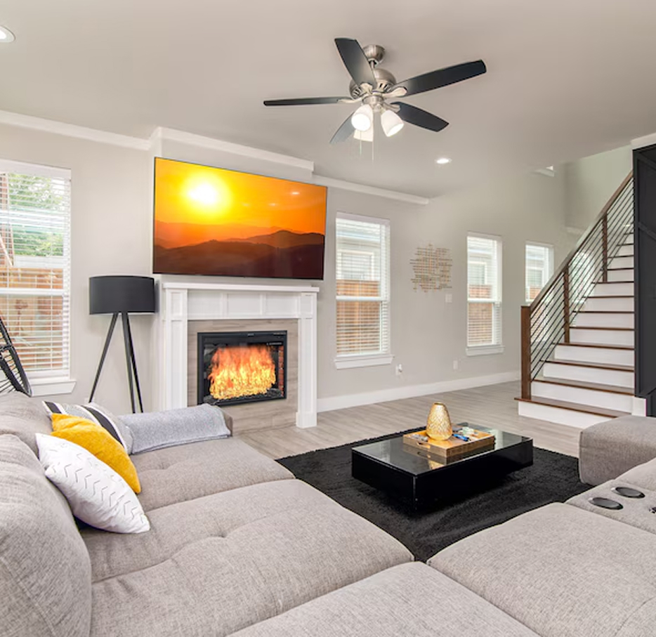 Modern living room with gray sectional, fireplace, wall-mounted TV, a black coffee table, and staircase.