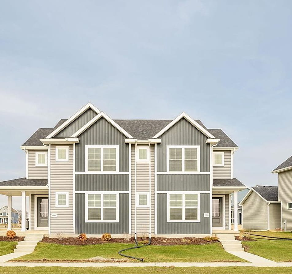 Symmetrical two-story duplex with gray siding, white trim, porches, and green lawn on a clear day.