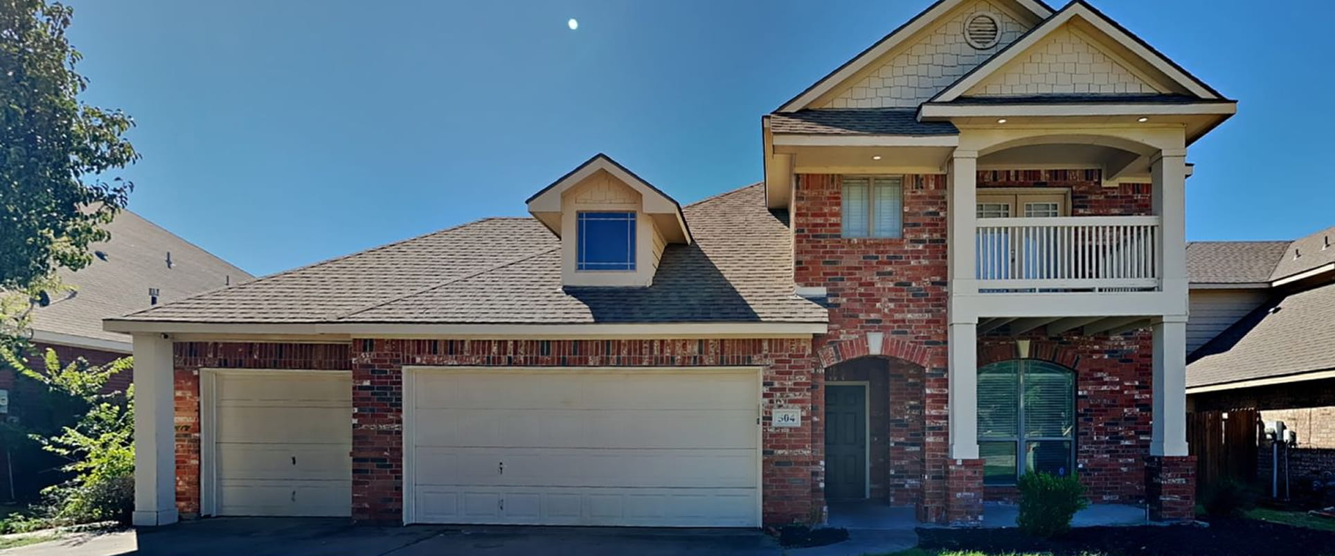 Two-story brick house with a double garage, a balcony, and a pitched roof under a clear blue sky.