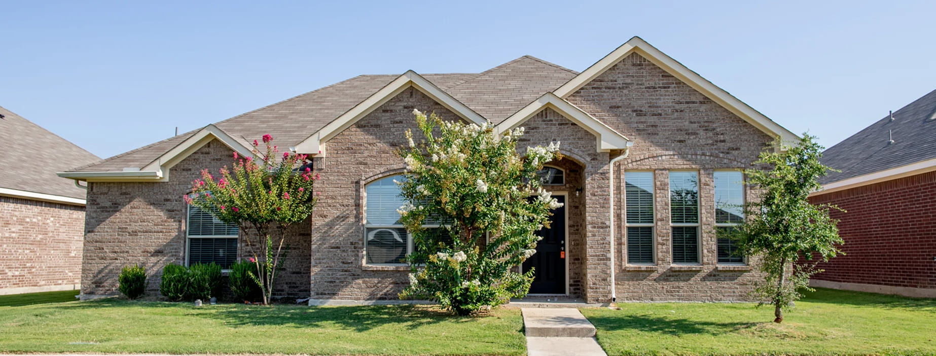 Single-story brick house with a black front door, multiple windows, and flowering trees in front yard.