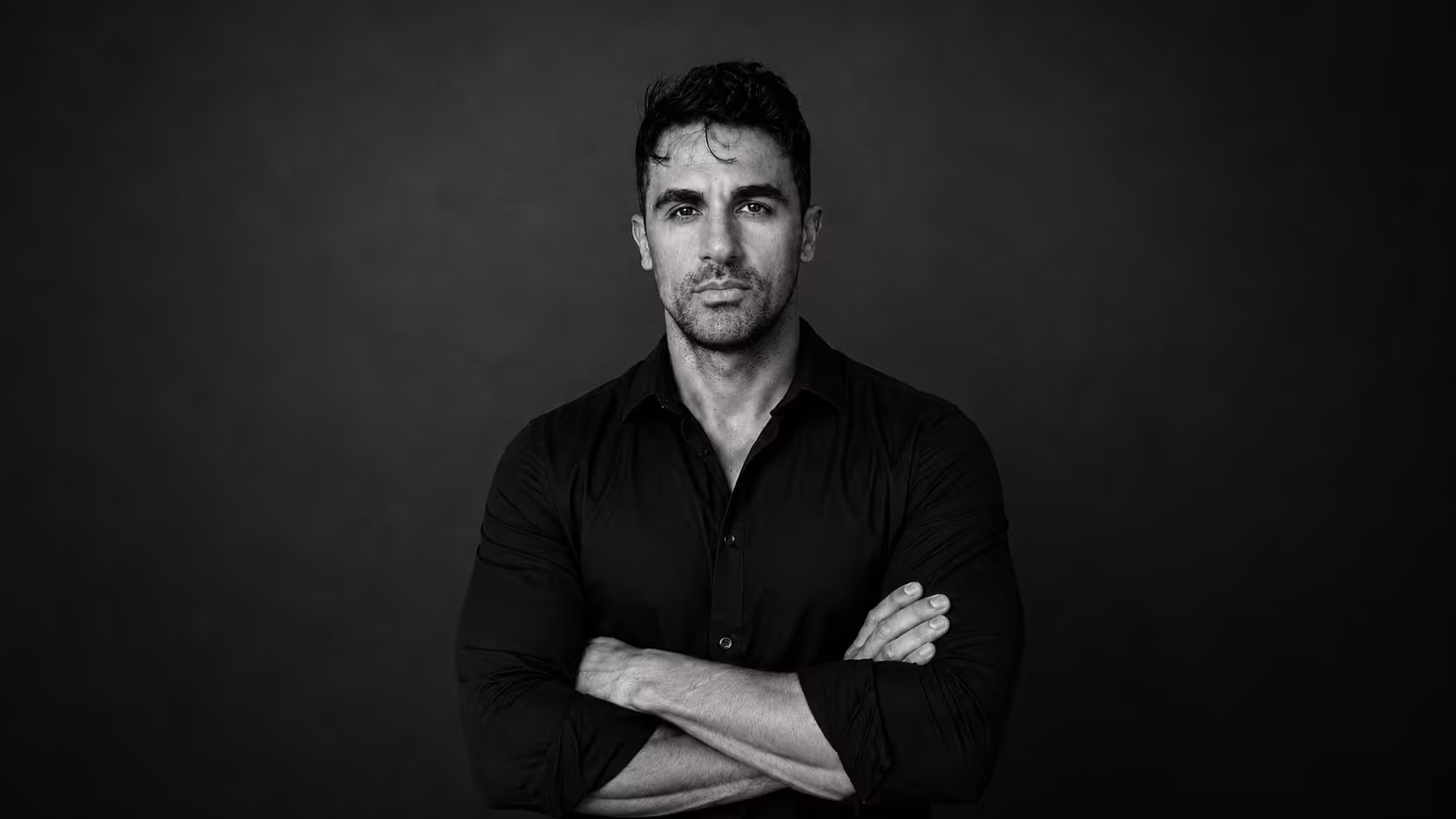 Serious man with dark hair and stubble in a black shirt, arms crossed, looking at the camera.