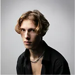 Young man with light wavy hair and necklace wearing a black shirt against a plain gray background.