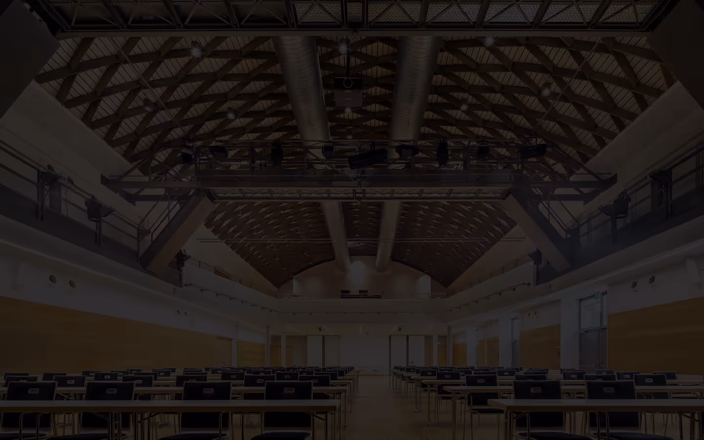 Large conference or lecture hall with rows of empty tables and chairs under a vaulted wooden ceiling with metal ductwork and overhead lighting rig.