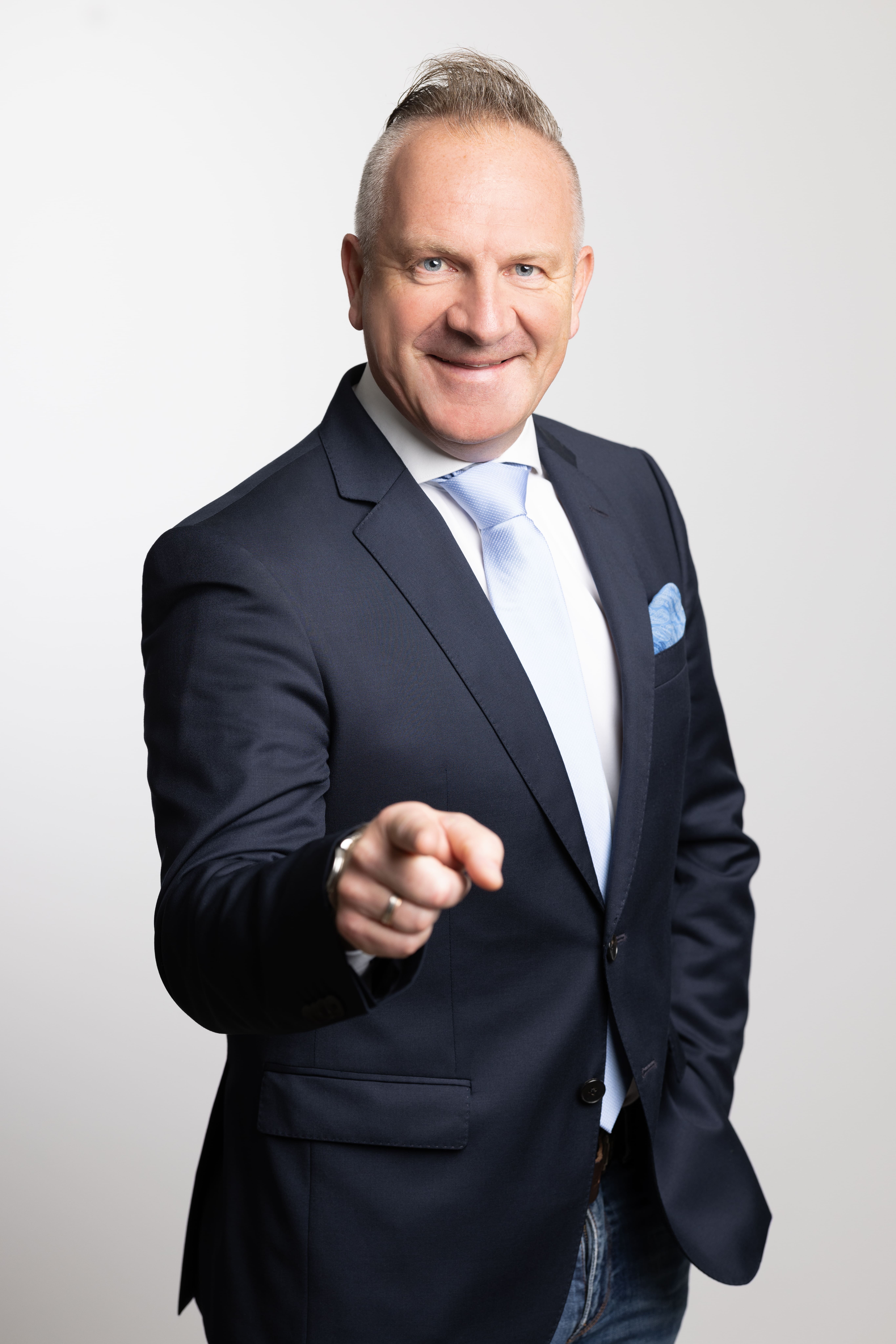 Lächelnder Mann im dunklen Anzug zeigt direkt auf die Kamera vor weißem Hintergrund.