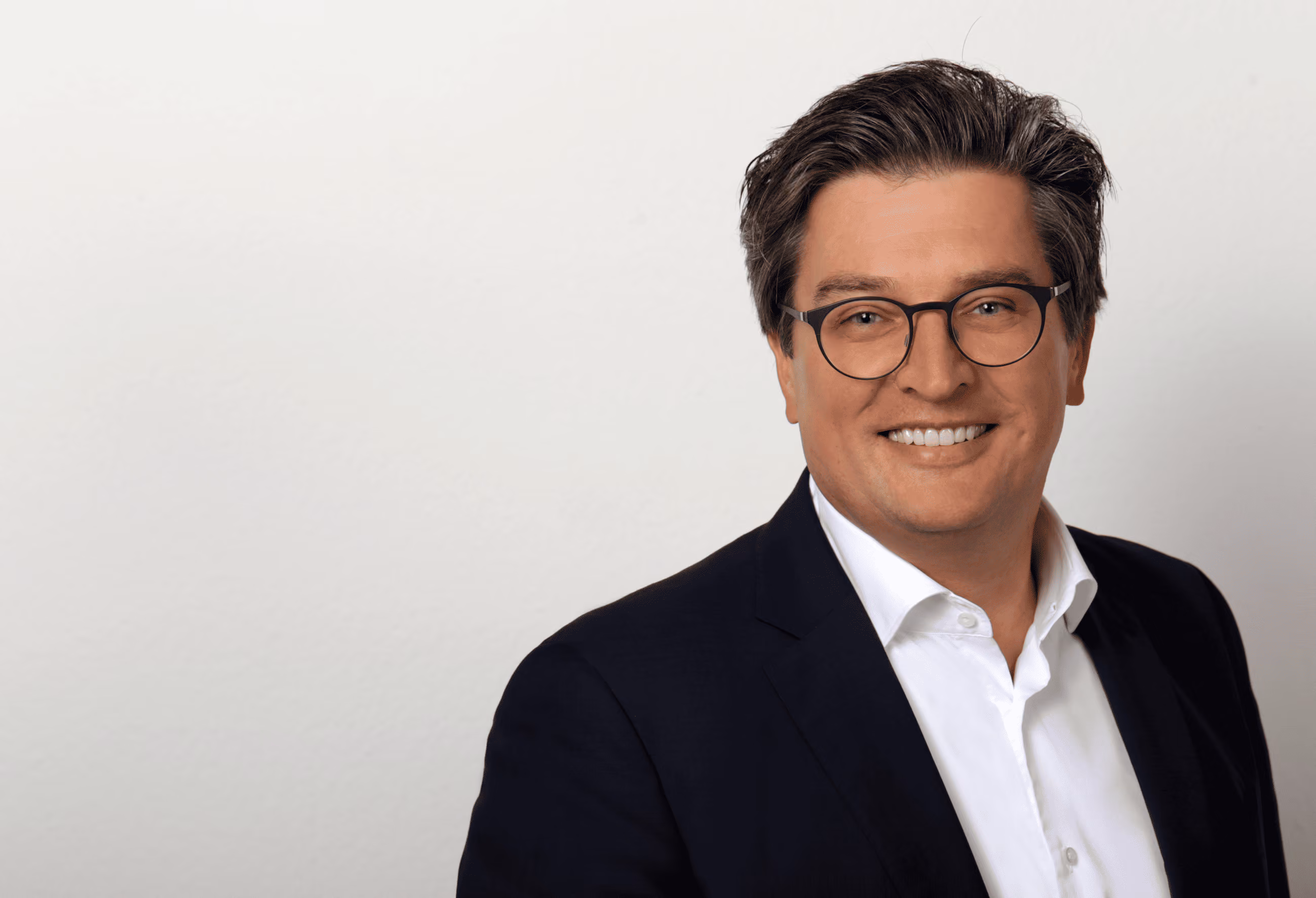 Mann mit Brille lächelt, trägt weißen Hemd und dunklen Anzug vor hellem Hintergrund.