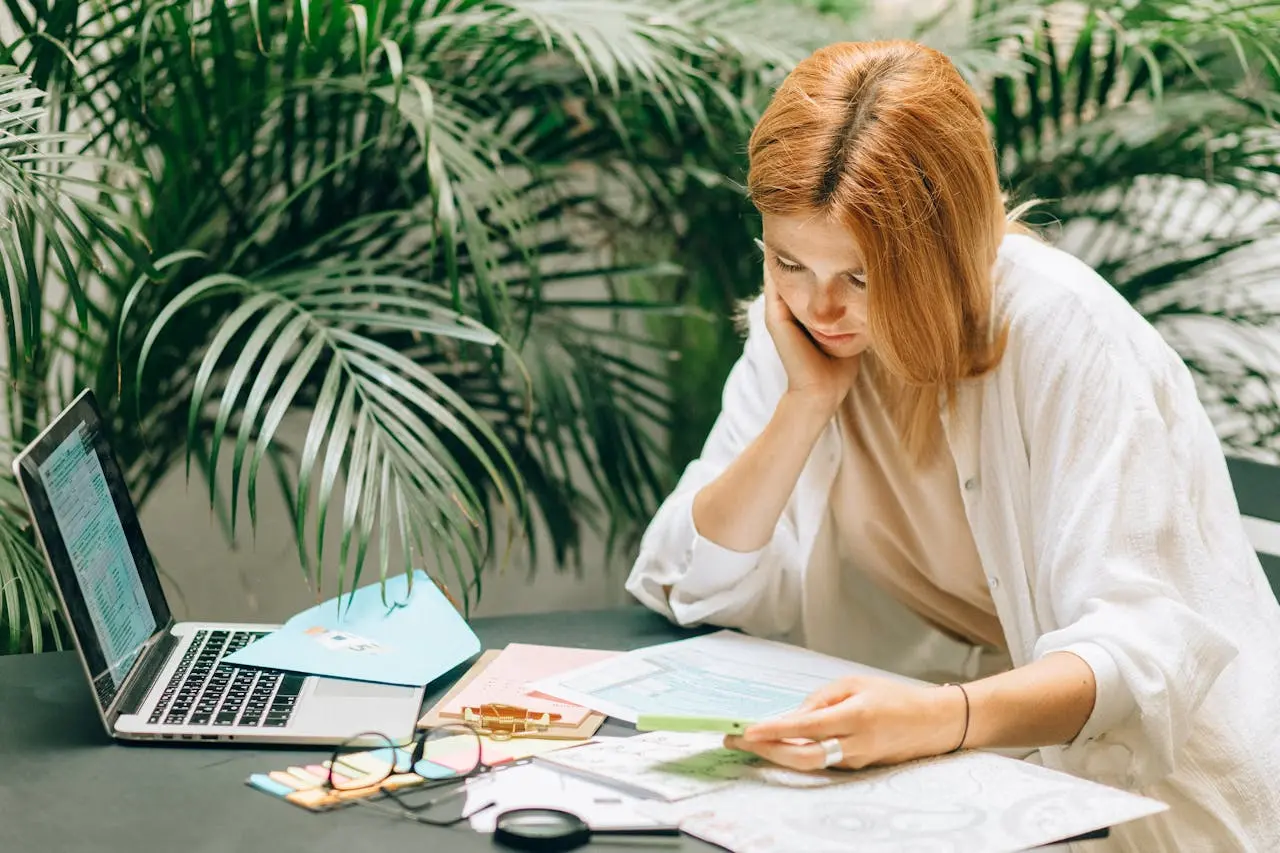 A woman sits at a table with documents, a laptop, notepad, and glasses, appearing to read or review papers in an indoor setting with green plants in the background.