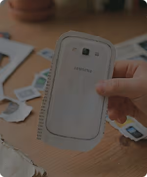 Hand holding a cutout of a Samsung smartphone back cover over a table with scattered cutouts.