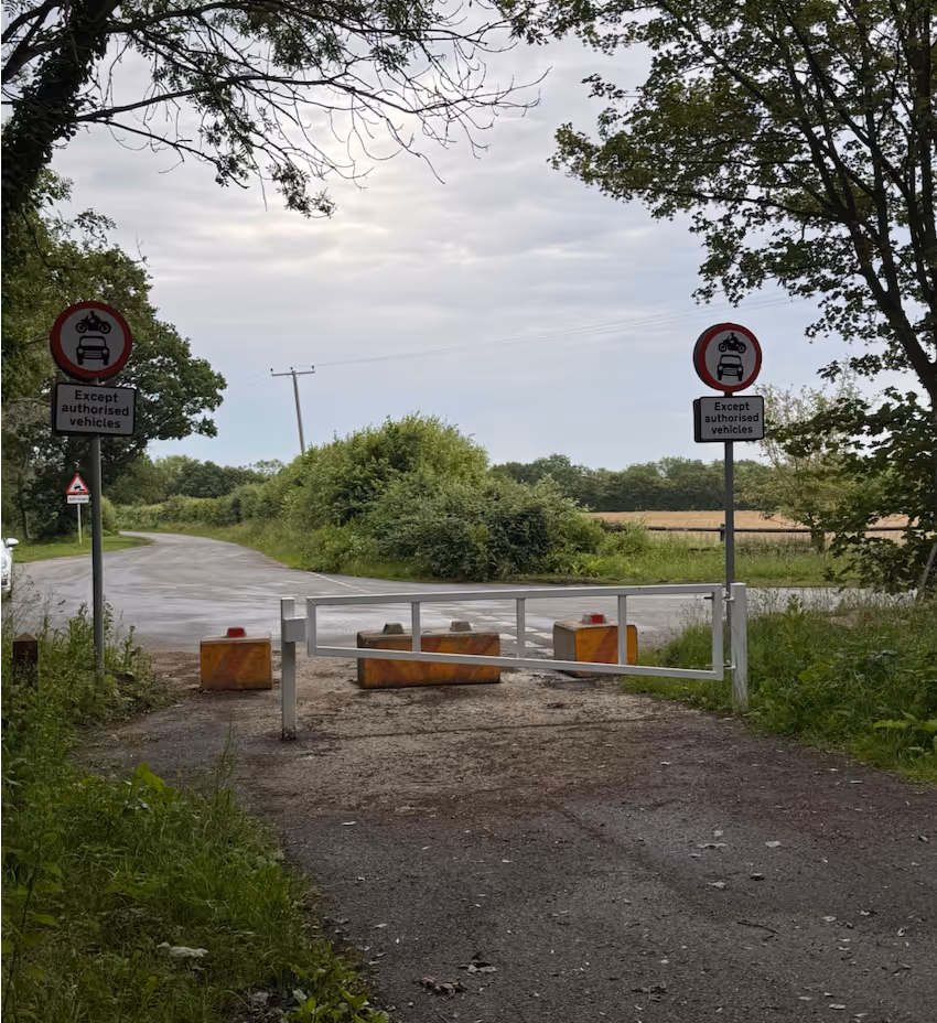 Rural road blocked by gate and concrete blocks with signs restricting vehicles except authorized ones.