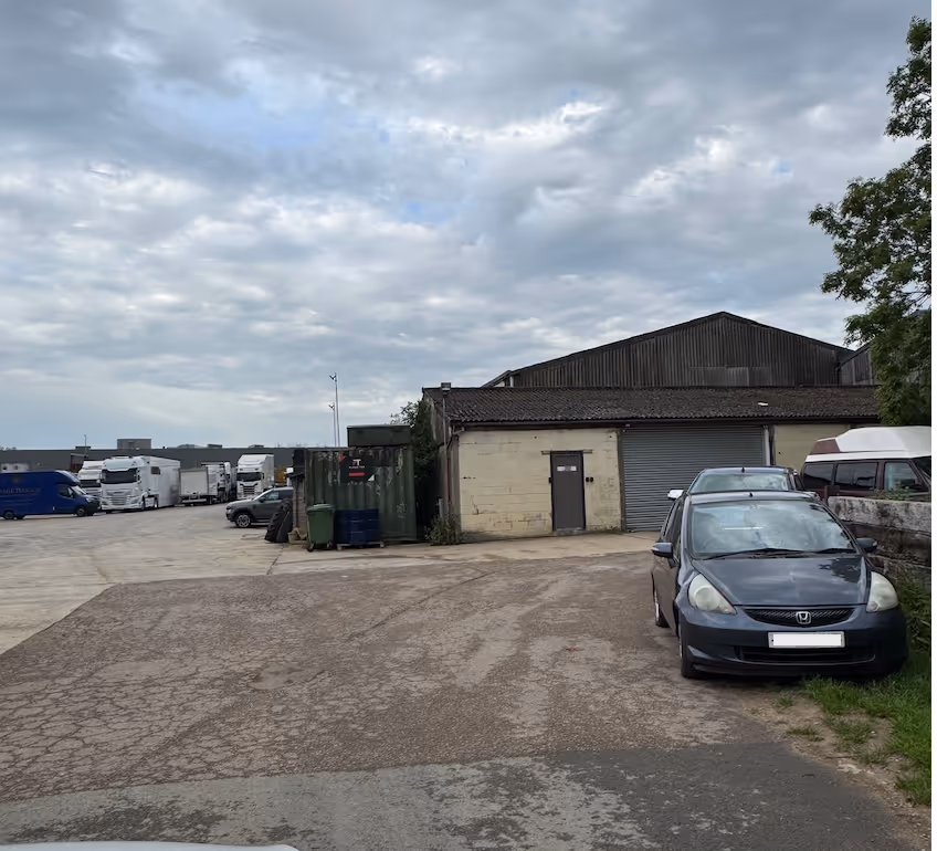 Industrial yard with parked trucks, a warehouse, and cars under a cloudy sky.