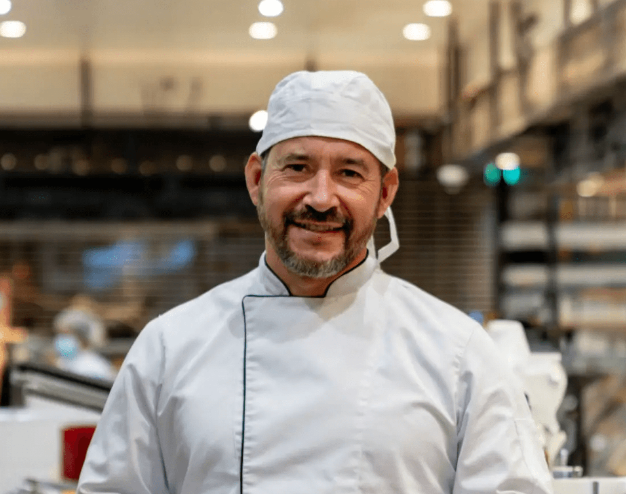 Smiling male chef wearing white uniform and cap in a bright kitchen setting.