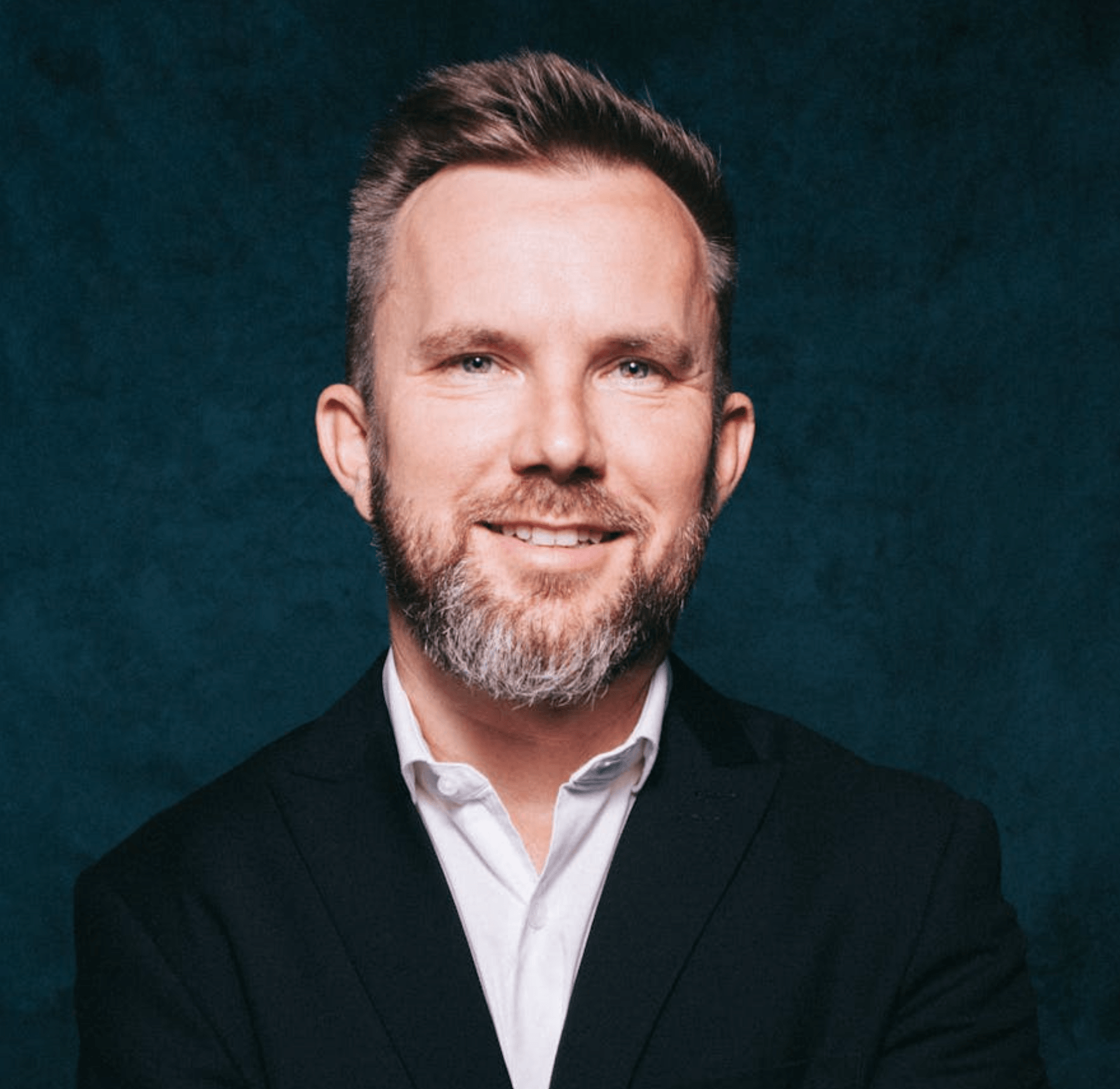 Smiling man with short hair and beard wearing a white shirt and dark suit jacket on a dark background.
