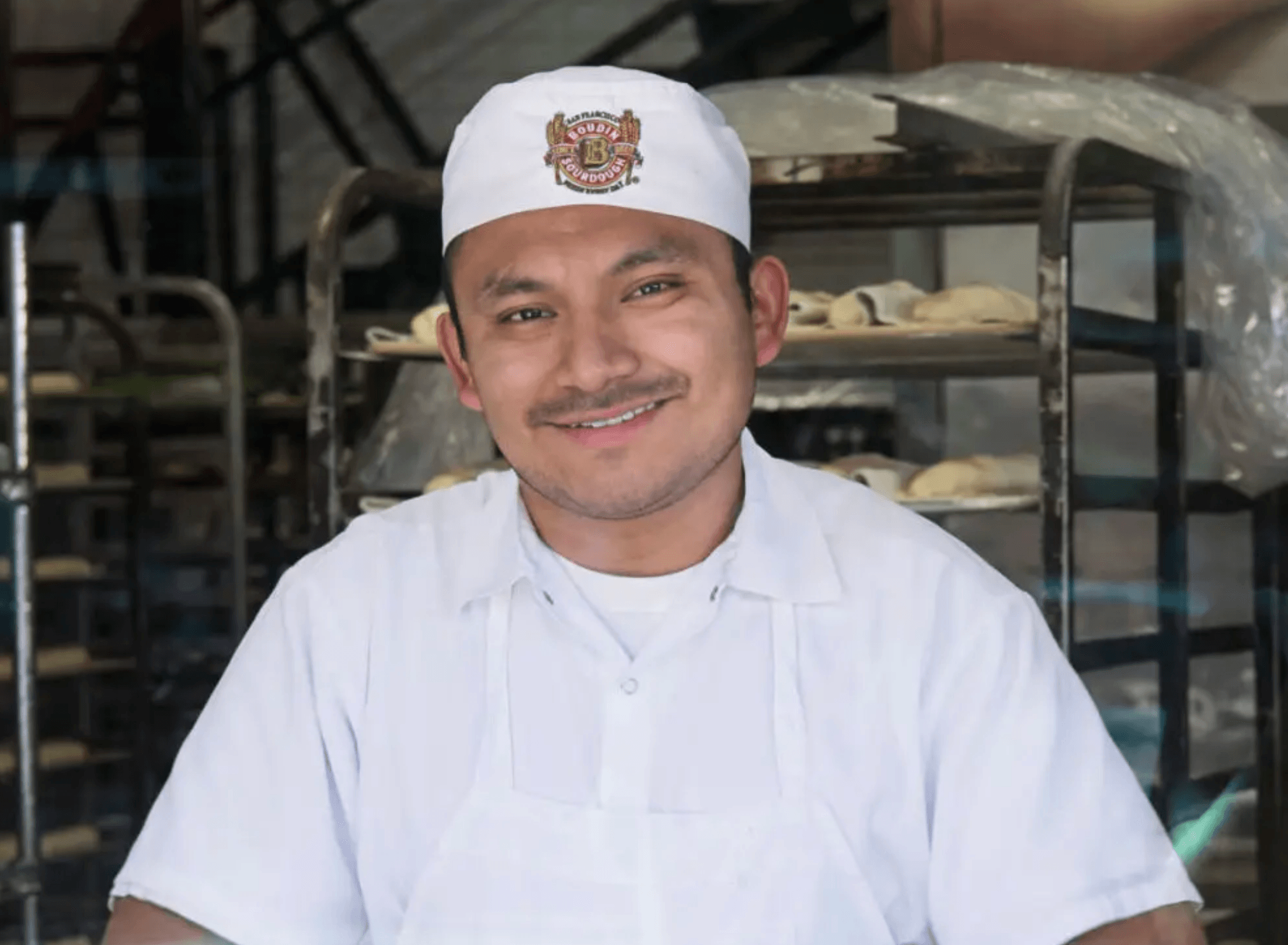 Smiling baker wearing white uniform and cap standing in front of bread trays in bakery kitchen.