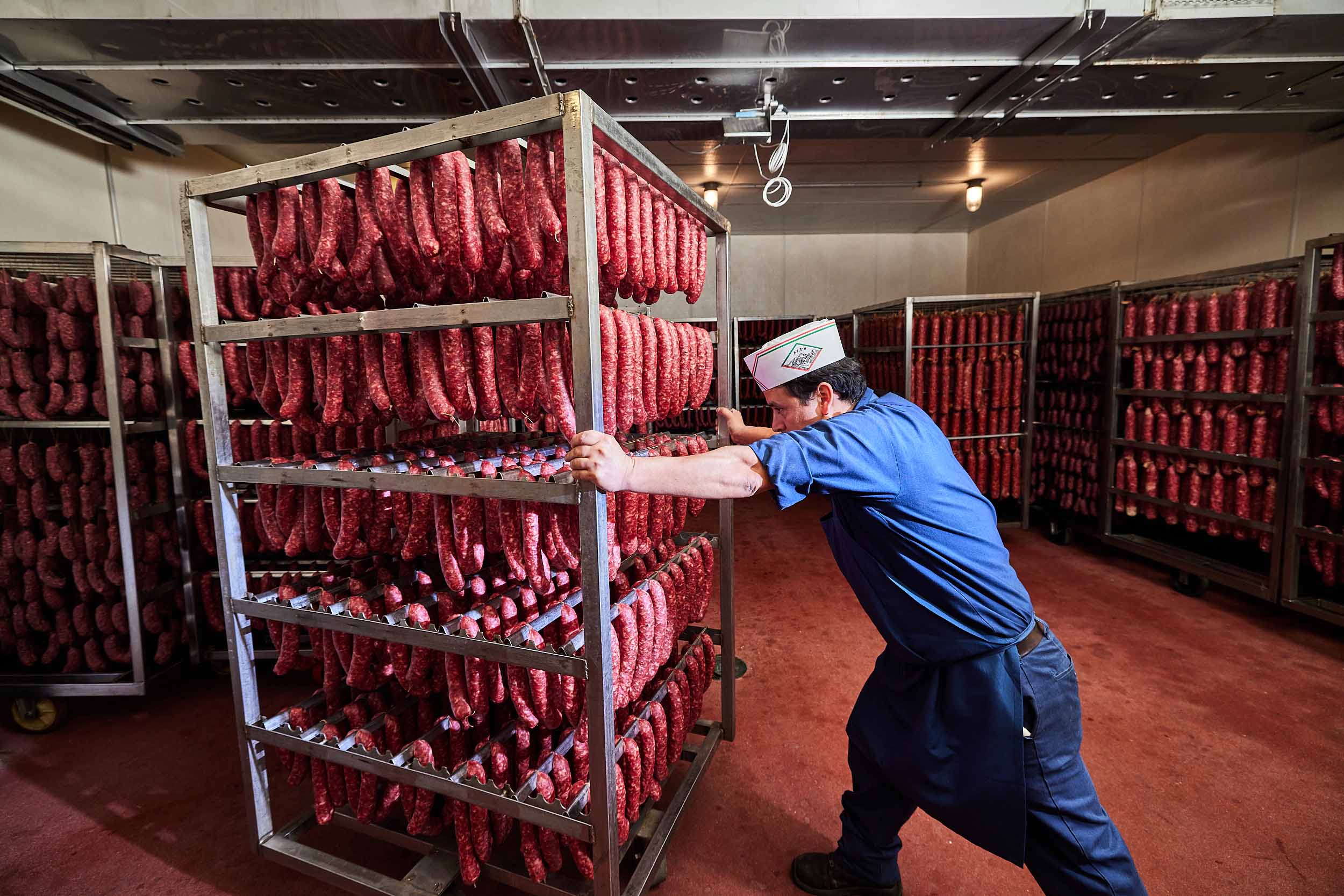 Worker in blue uniform pushing rack filled with rows of hanging sausages in meat processing plant.