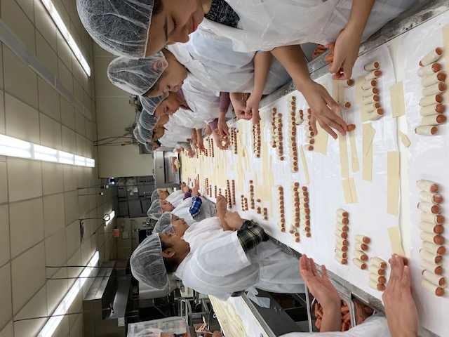 Workers in hairnets and aprons assembling rows of small wrapped sausages on a long table in a factory kitchen.