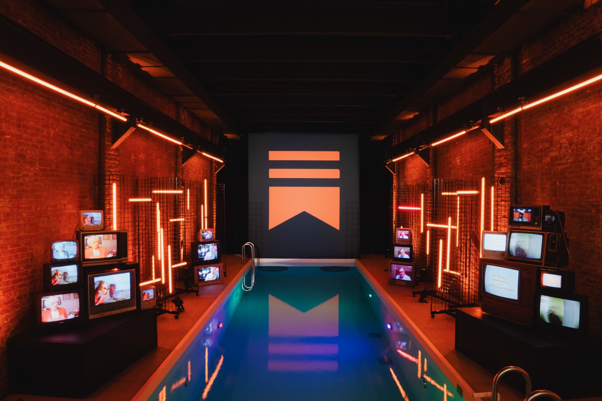 Industrial event venue with exposed brick walls lit by orange neon tubes, stacks of vintage TV monitors, and a swimming pool reflecting the warm lighting
