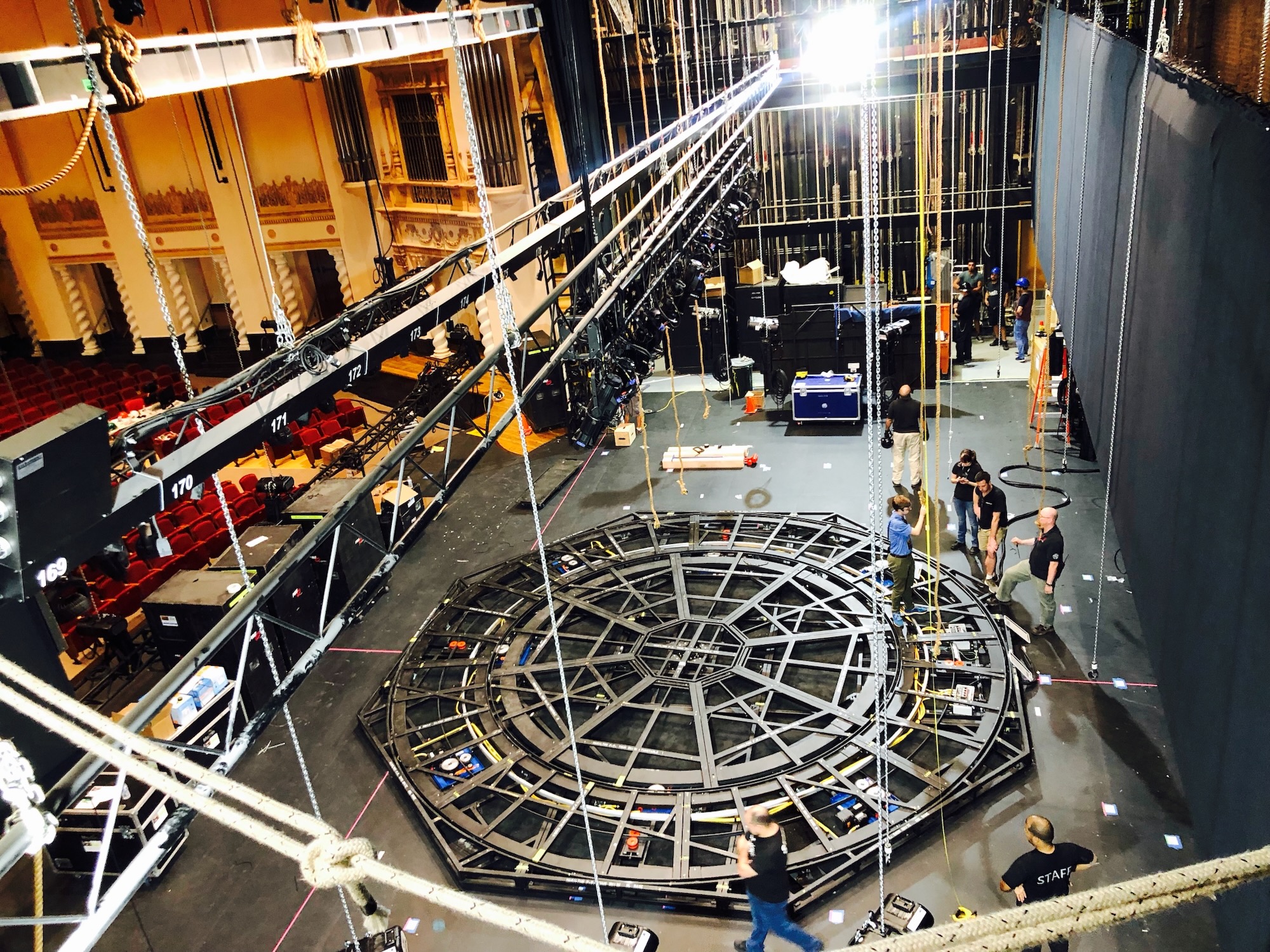 Backstage load-in at the theater: the circular Hamilton turntable being assembled on the stage deck by the production crew, with rigging lines and flight cases during the build.