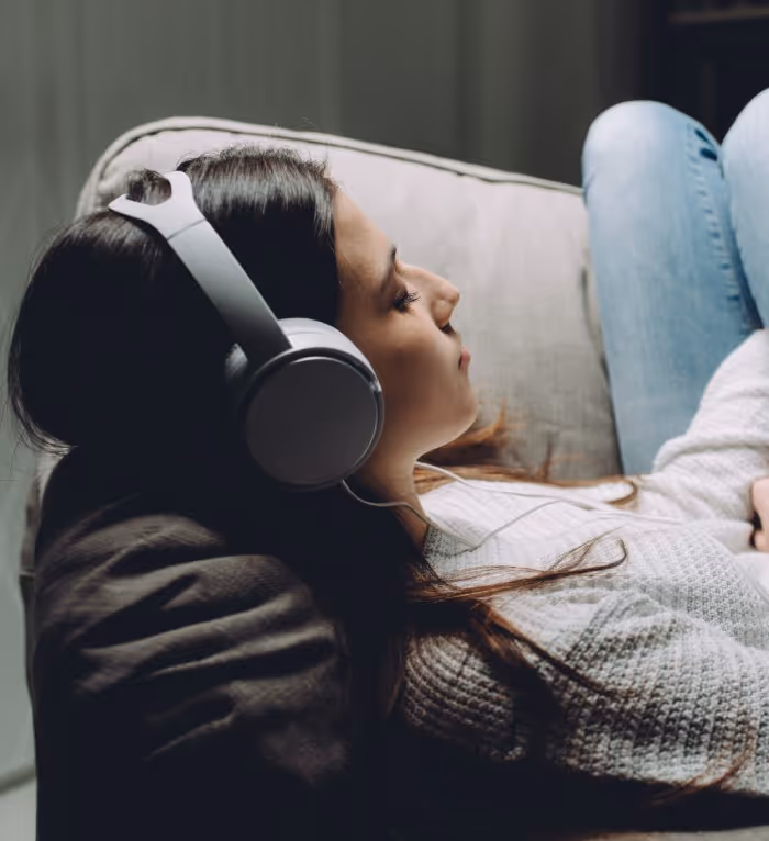 Woman reclining on couch with closed eyes wearing white headphones and blue jeans.