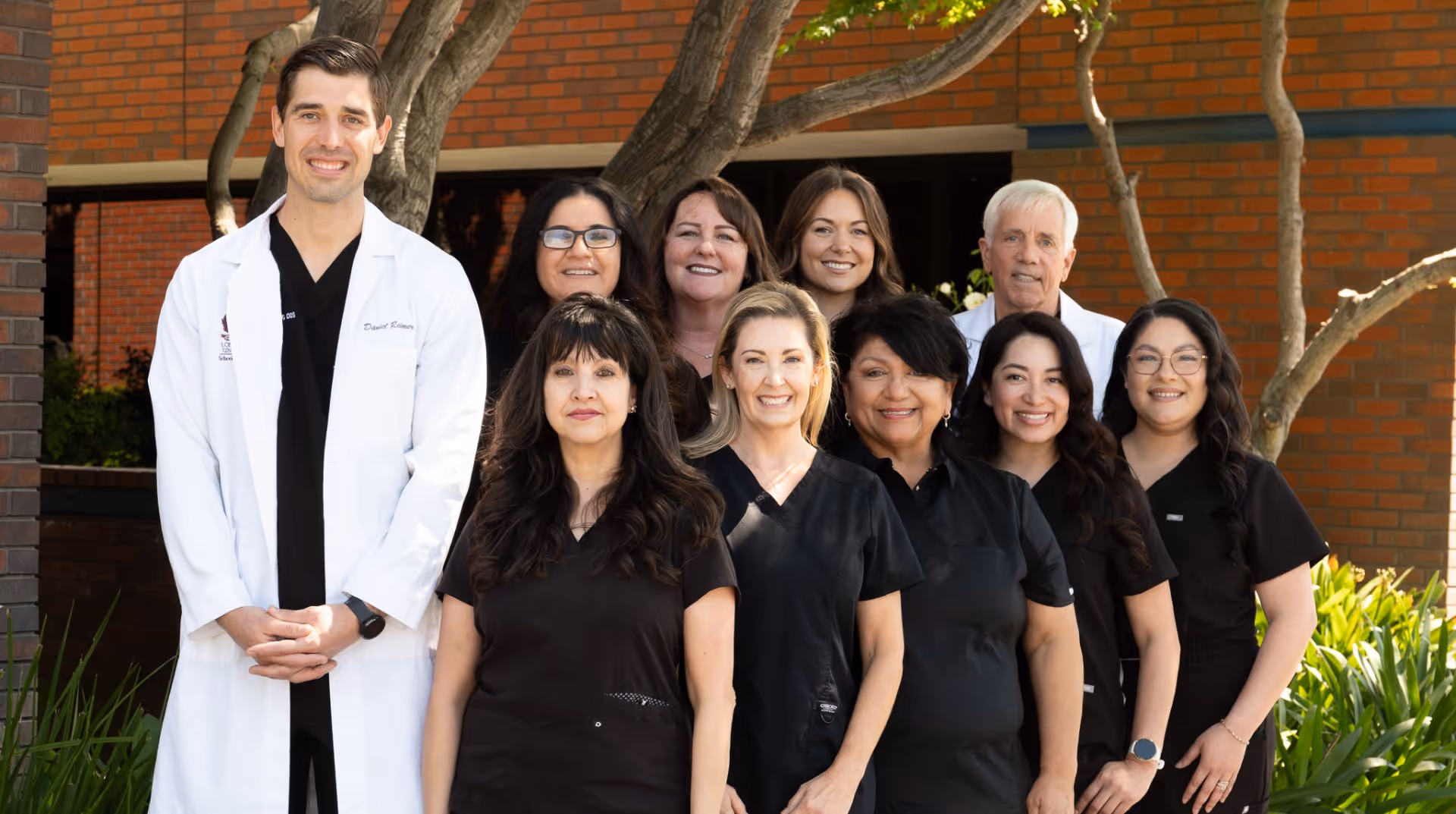 Smiling dental team of nine in black scrubs and two in white coats outside a brick building.