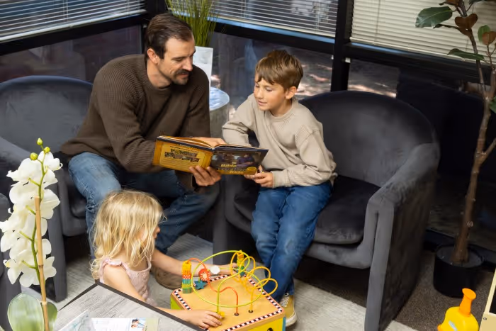 Man reads a book to a boy seated on a chair while a girl plays with a bead maze toy nearby.