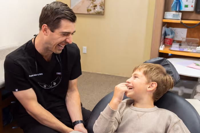 Dentist in black scrubs smiling and talking to a young boy sitting in a dental chair.