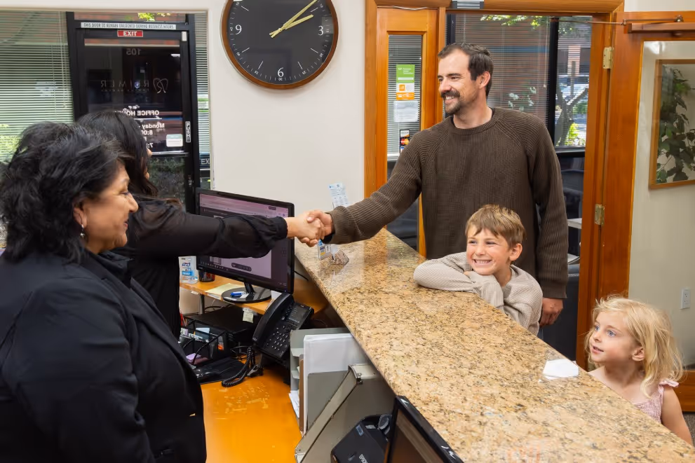 Man shaking hands with reception worker while two children watch across office counter.