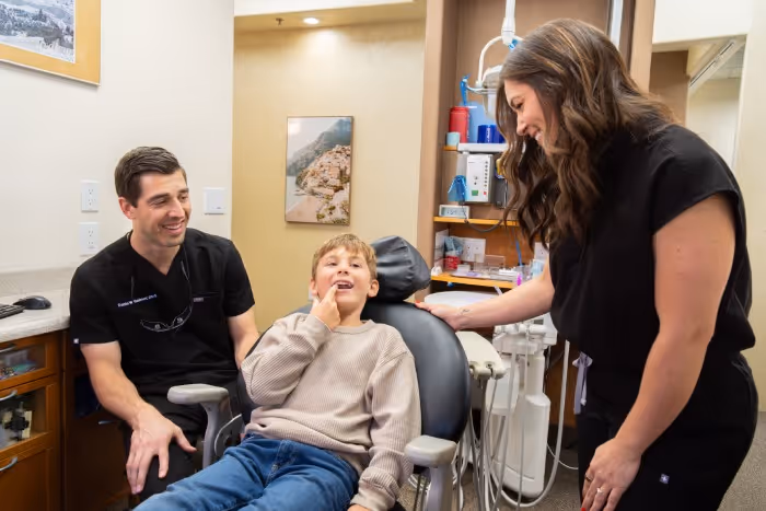 Smiling dentist and dental assistant talk to a boy sitting in a dental chair in clinic room.
