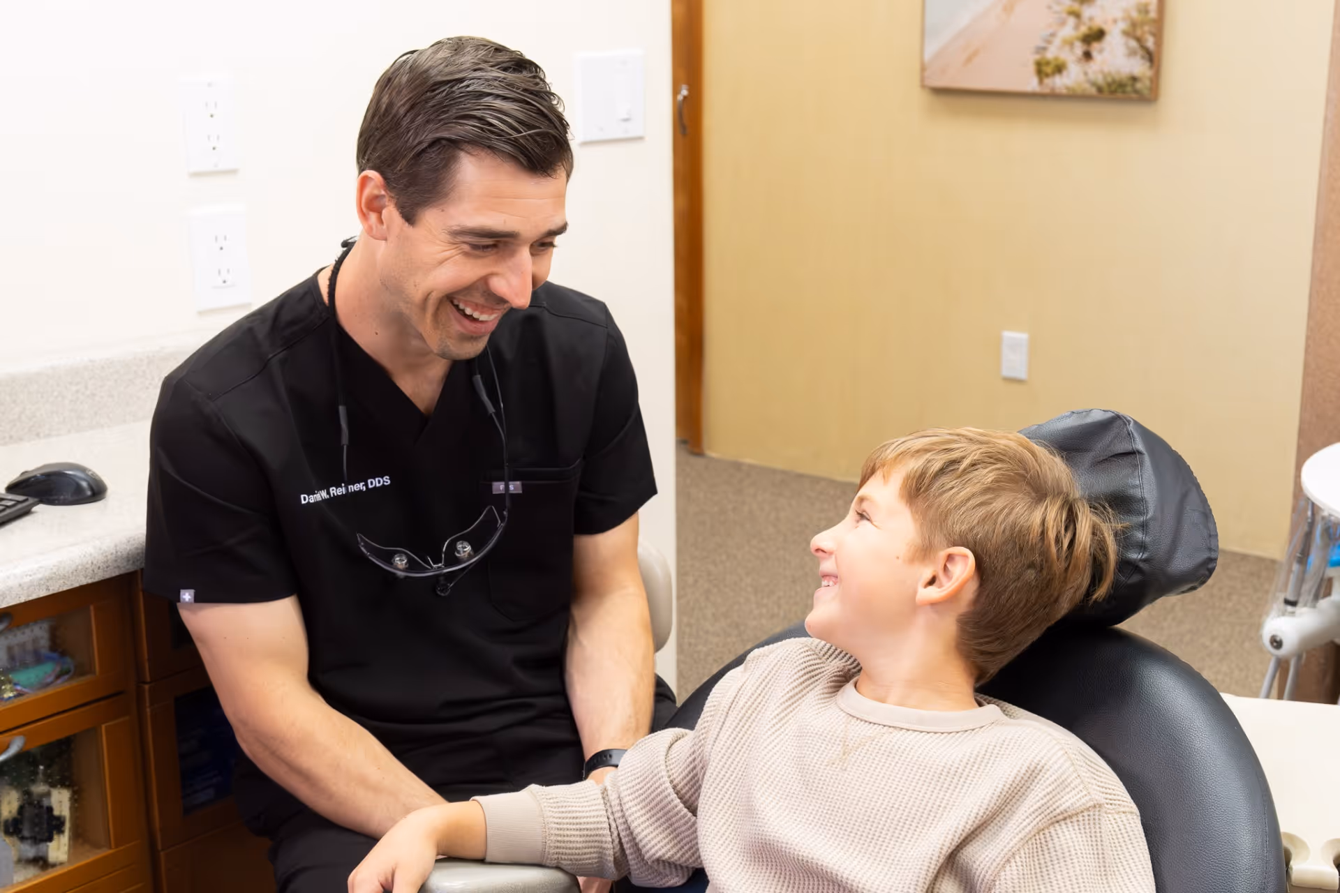 Smiling male dentist in black scrubs talks with a relaxed boy in a dental chair.