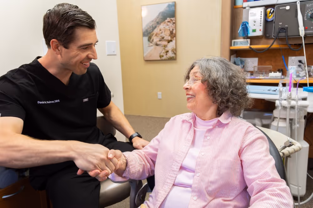 Dentist in black scrubs shaking hands and smiling with an elderly female patient in the office.