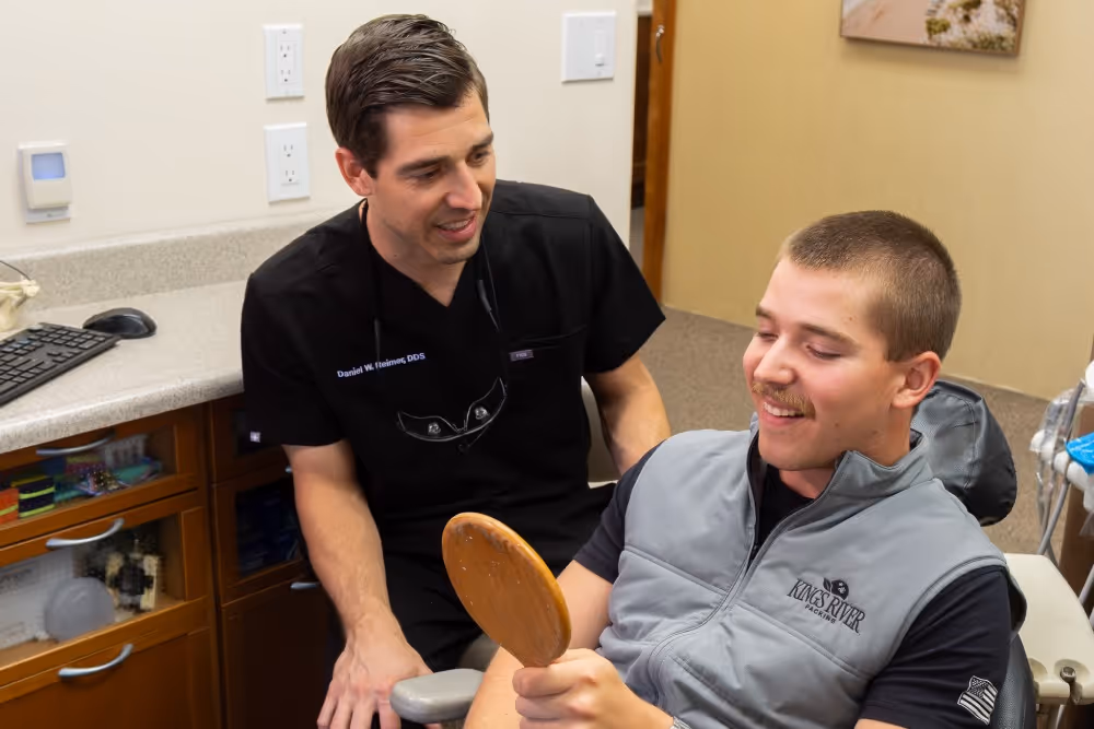 Male dentist in black scrubs talks to smiling male patient holding a hand mirror in dental office.