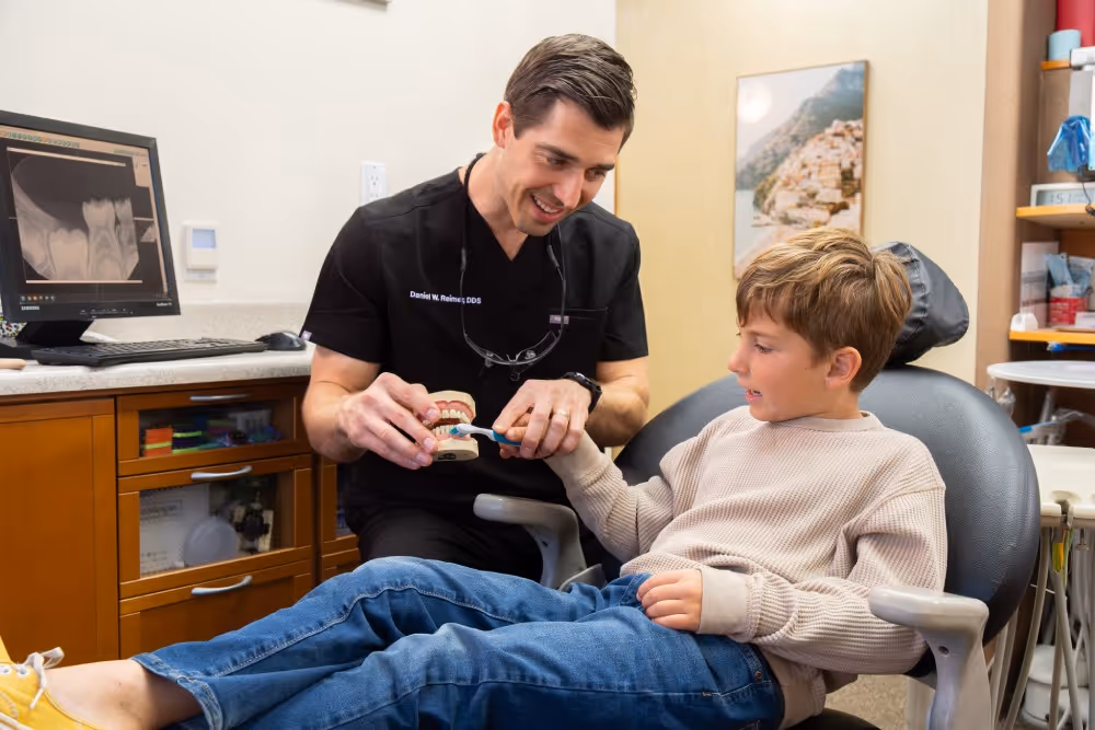 Dentist in black scrubs teaching a boy how to brush teeth with a model in a dental office.