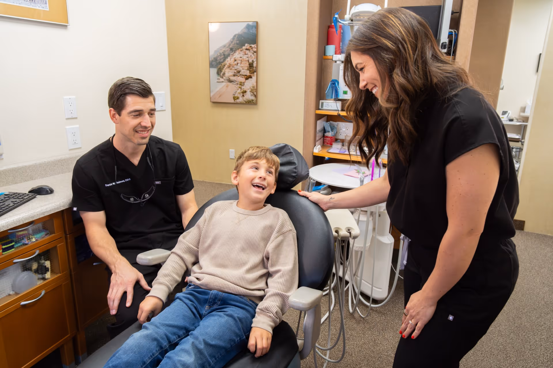Boy sitting in a dental chair smiling at a woman while a male dentist watches nearby in an office.