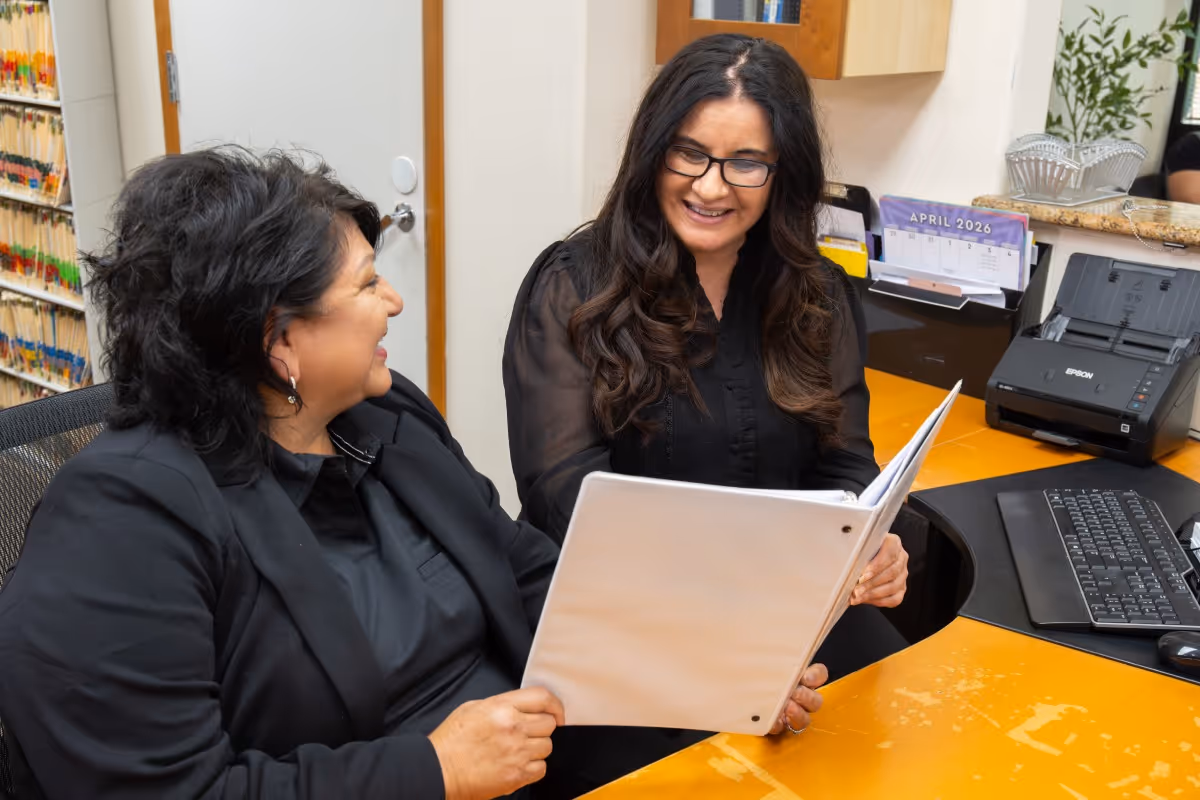 Two women smiling and looking at a binder while sitting at an office desk with files and a calendar.