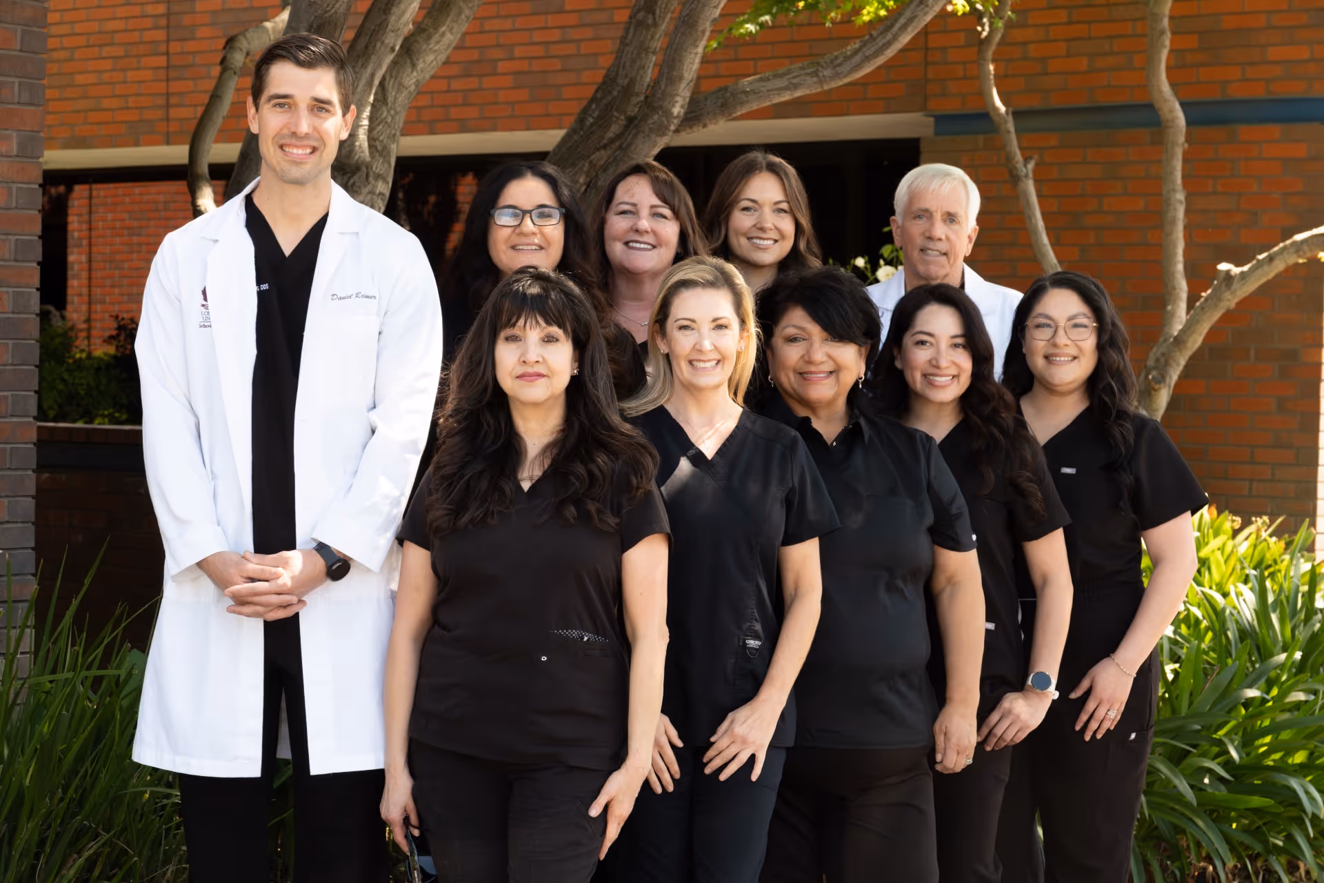 Group of ten smiling healthcare professionals posing outdoors in medical uniforms and a white coat.