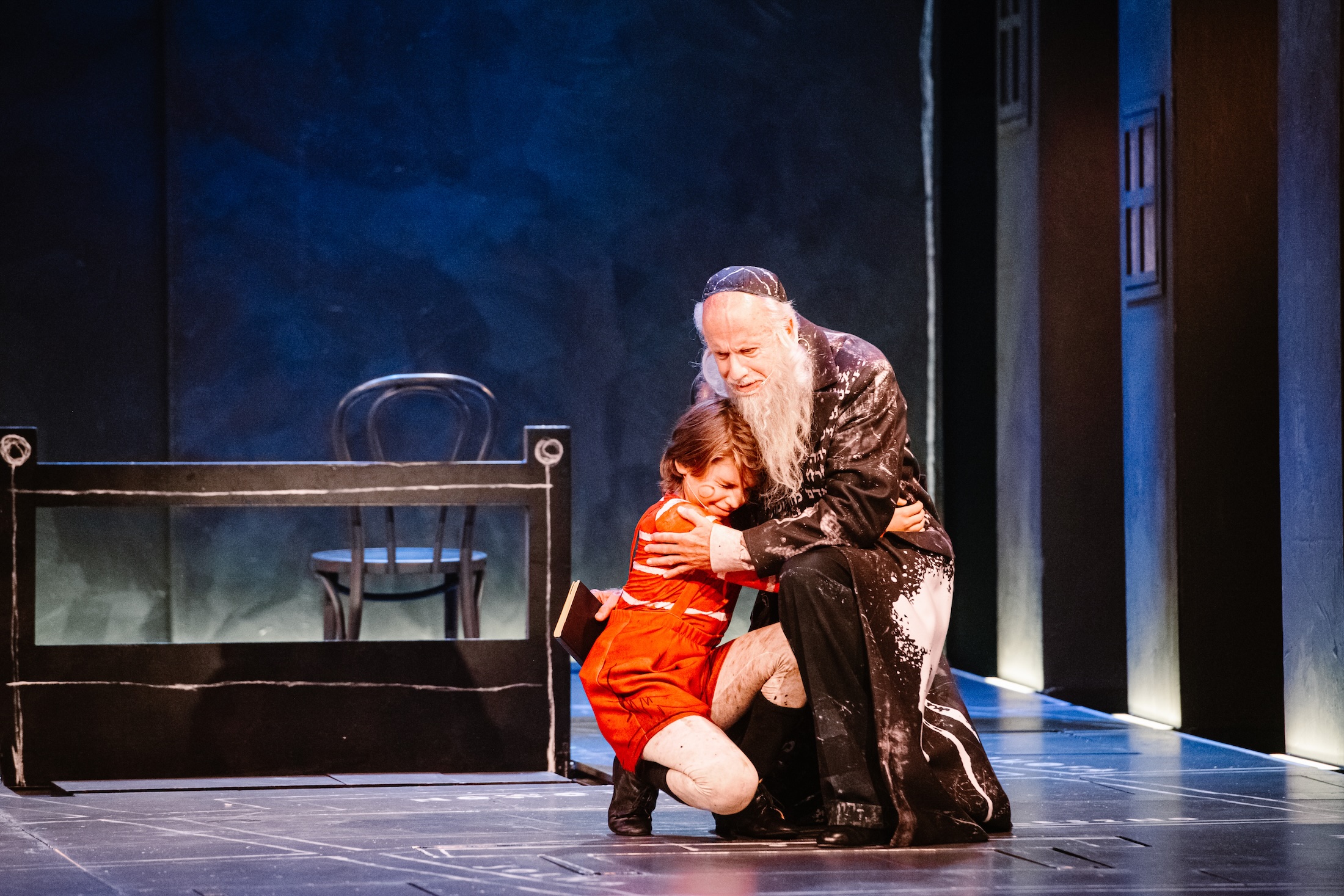 An older man embraces a young boy in red overalls onstage in Irena.
