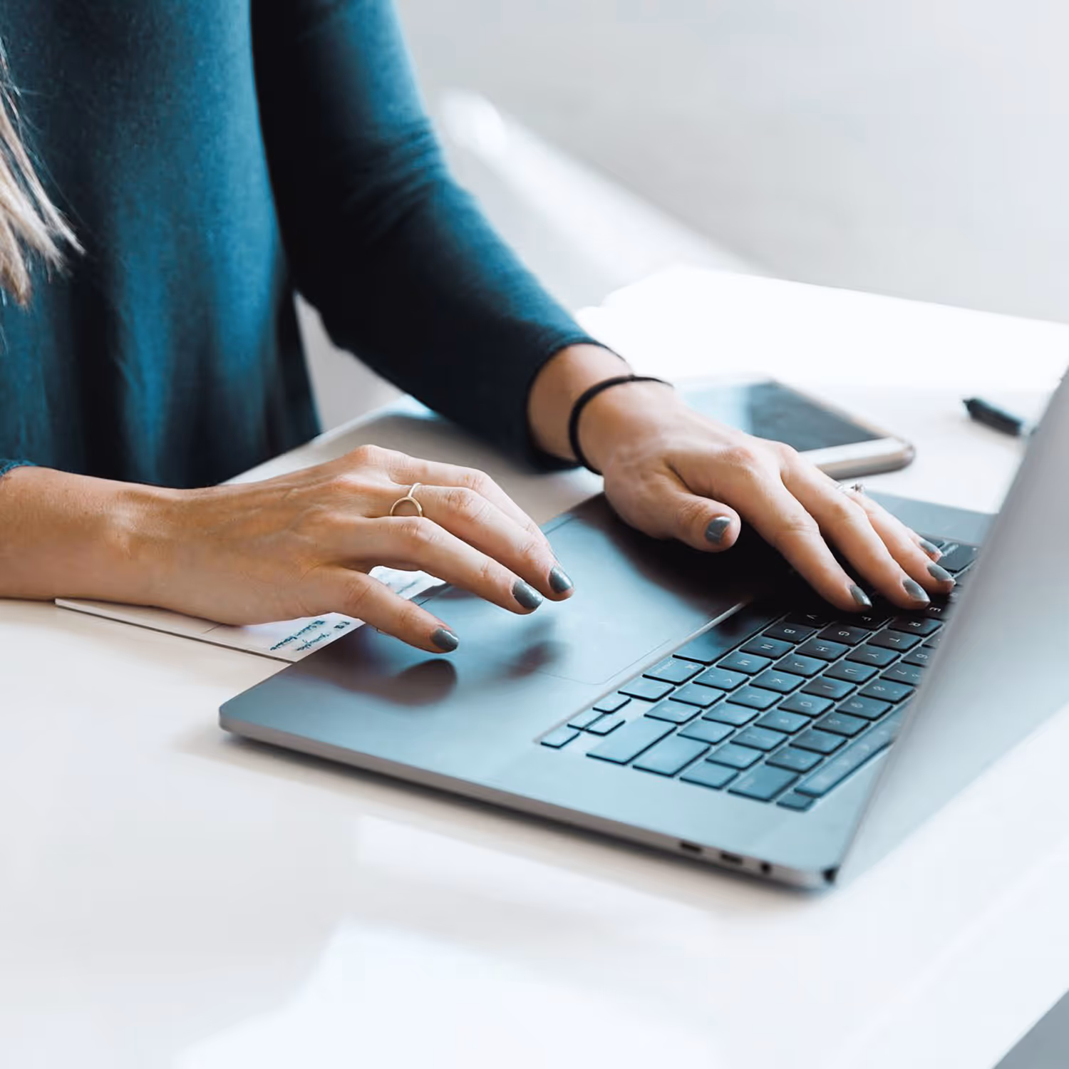 Person with painted nails typing on a laptop keyboard at a white desk with a smartphone nearby.
