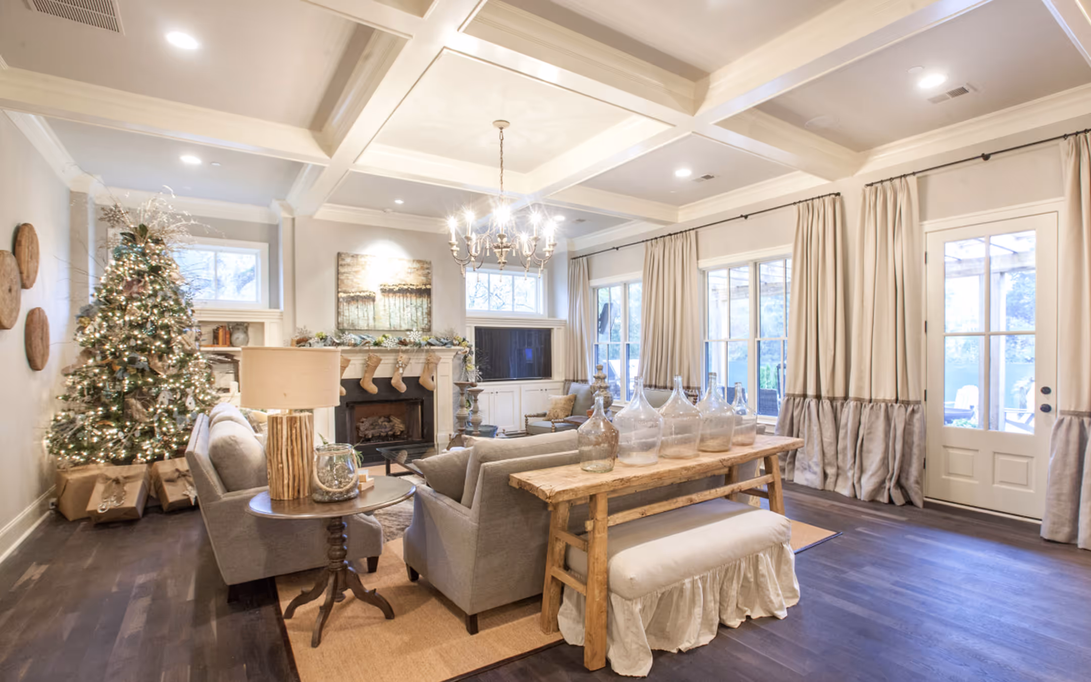 Cozy living room with a decorated Christmas tree, gray sofas, fireplace, and large windows with curtains.