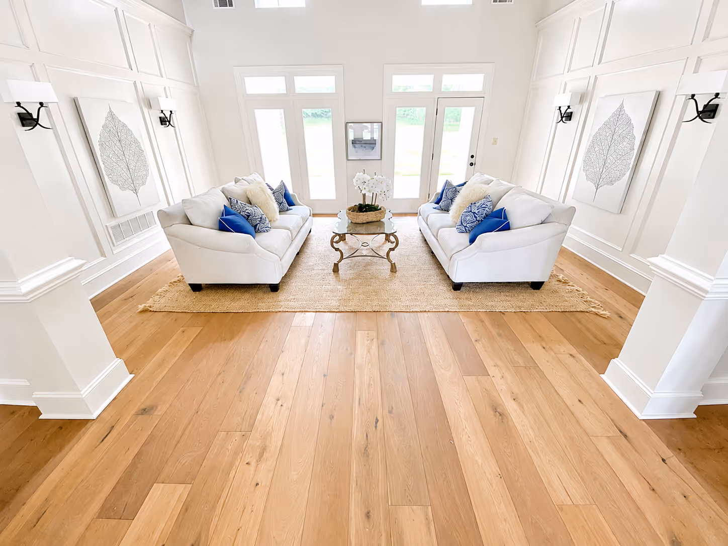 Bright living room with two white sofas, blue and white pillows, glass coffee table, and wooden floor.