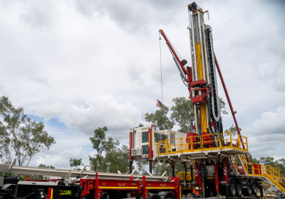 A large red and yellow drilling rig is set up outdoors under a cloudy sky, with the mast fully raised and support equipment positioned around the base. The rig sits among sparse trees, with safety railings and operational platforms visible, highlighting a 