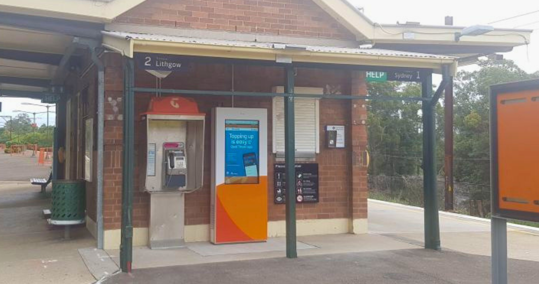 Train station platform showing a brick shelter with a public payphone, an orange ticket vending machine, and platform signs indicating directions to Lithgow on platform 2 and Sydney on platform 1, with a HELP sign visible.