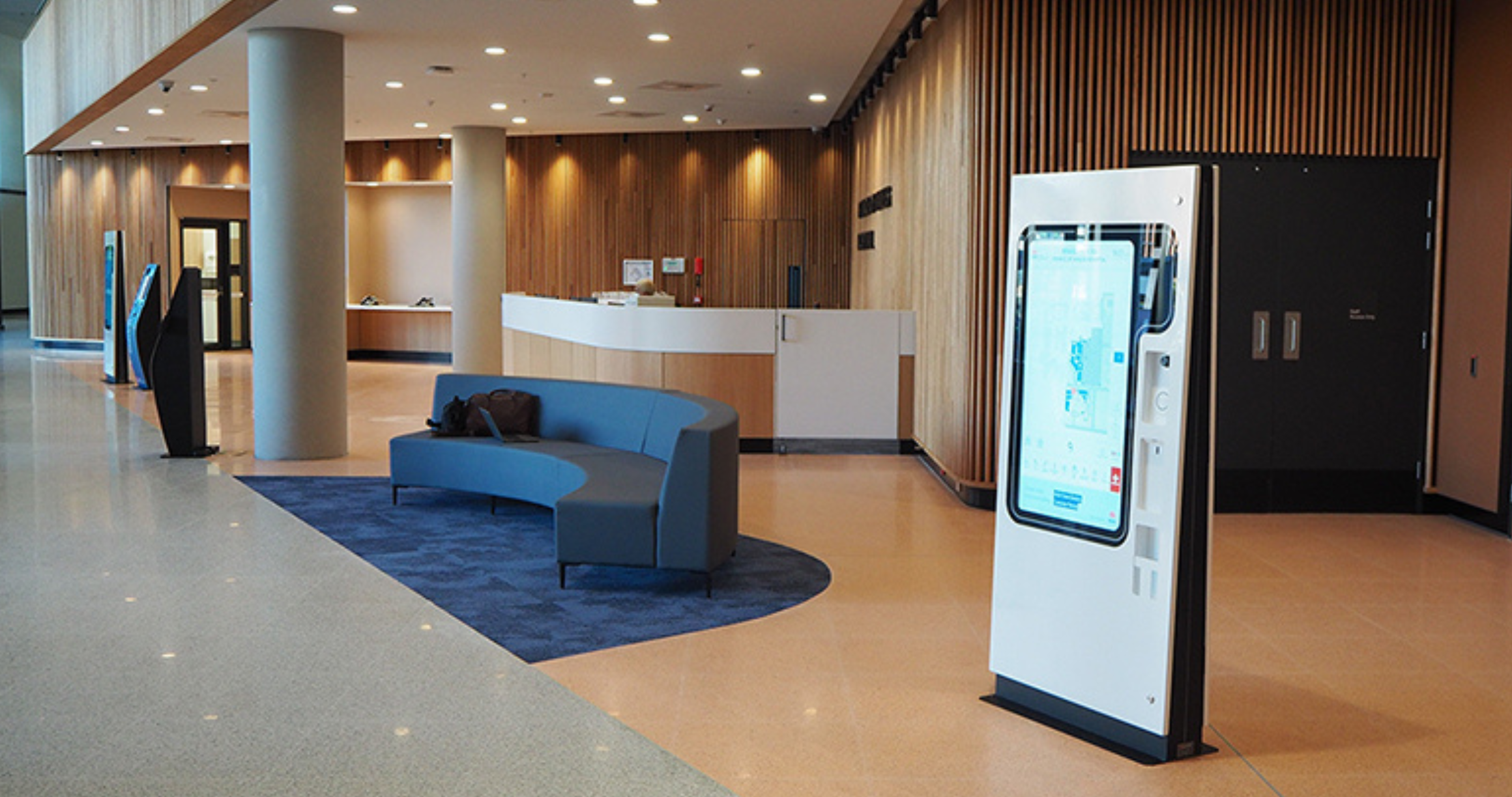 Modern lobby area with a curved blue seating arrangement on a blue rug, a white reception desk in the background, wooden paneled walls, and a large freestanding digital kiosk with a touchscreen display on the right side.