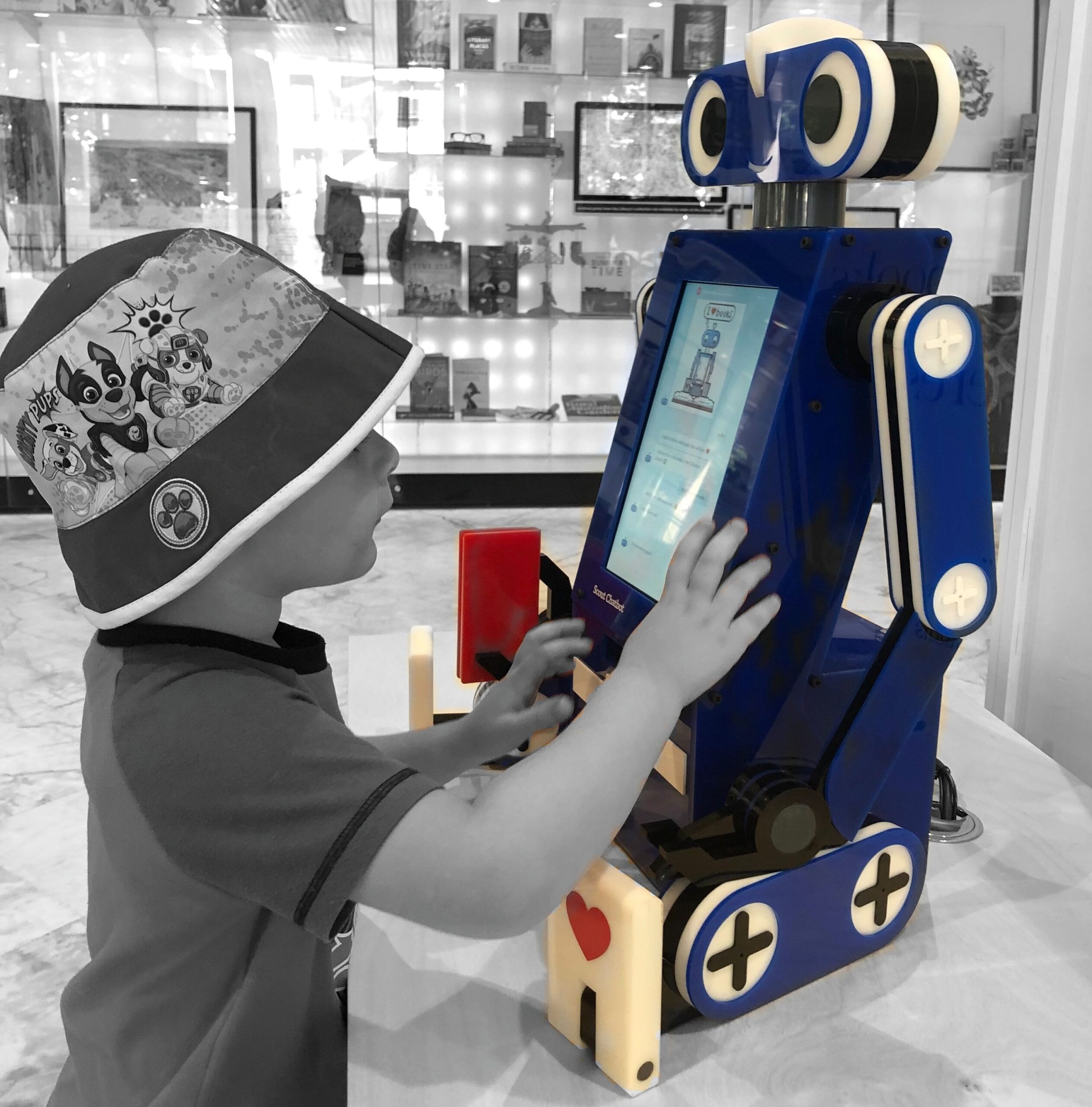A child wearing a Paw Patrol hat interacts with a blue robot that has a touchscreen on its chest, mounted on a table in a room with shelves and framed pictures in the background.