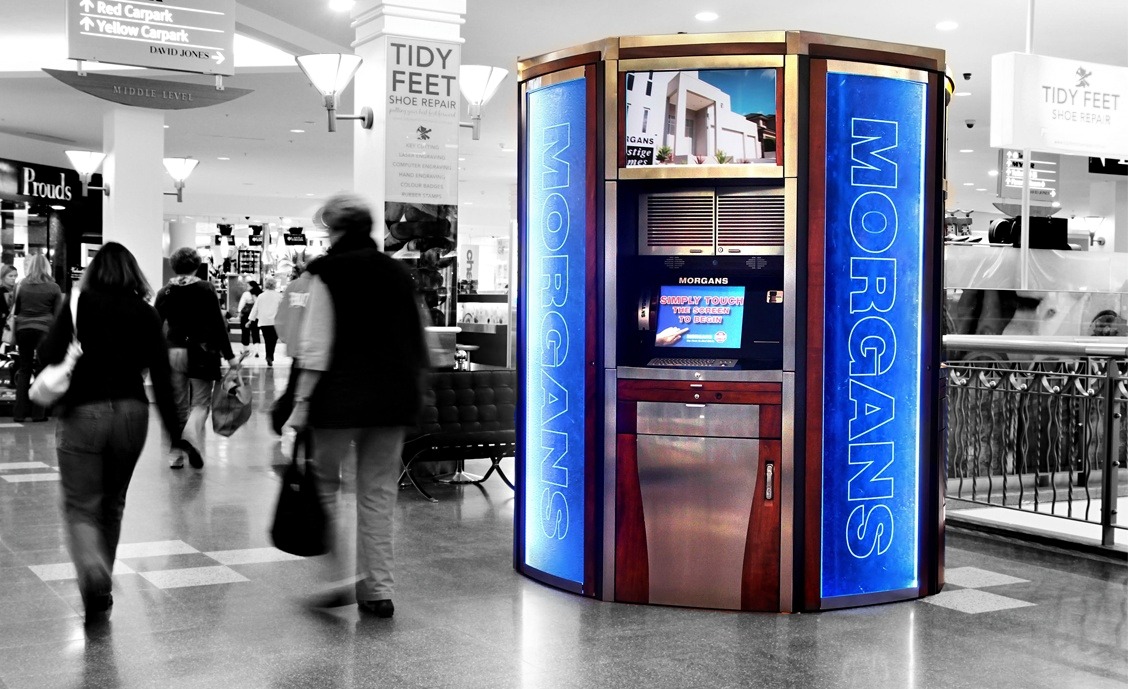 An ATM kiosk with blue illuminated panels displaying the name 'MORGANS' in a shopping mall corridor, with blurred people walking by and signs for Tidy Feet shoe repair and carparks overhead.