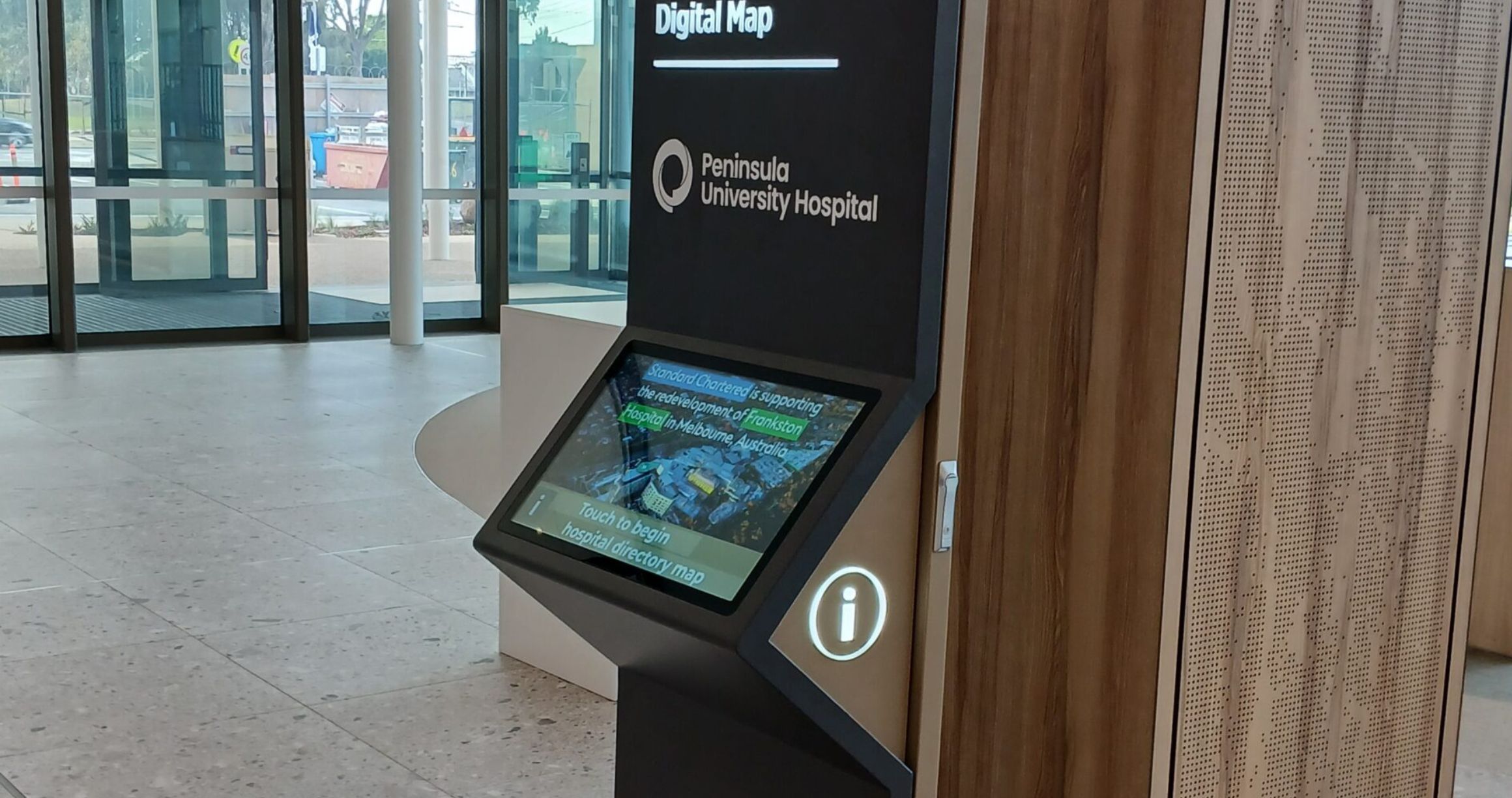 Digital wayfinding kiosk inside a building with a touchscreen displaying a map and text about the redevelopment of Frankston Hospital in Melbourne, Australia, and the Peninsula University Hospital logo above it.