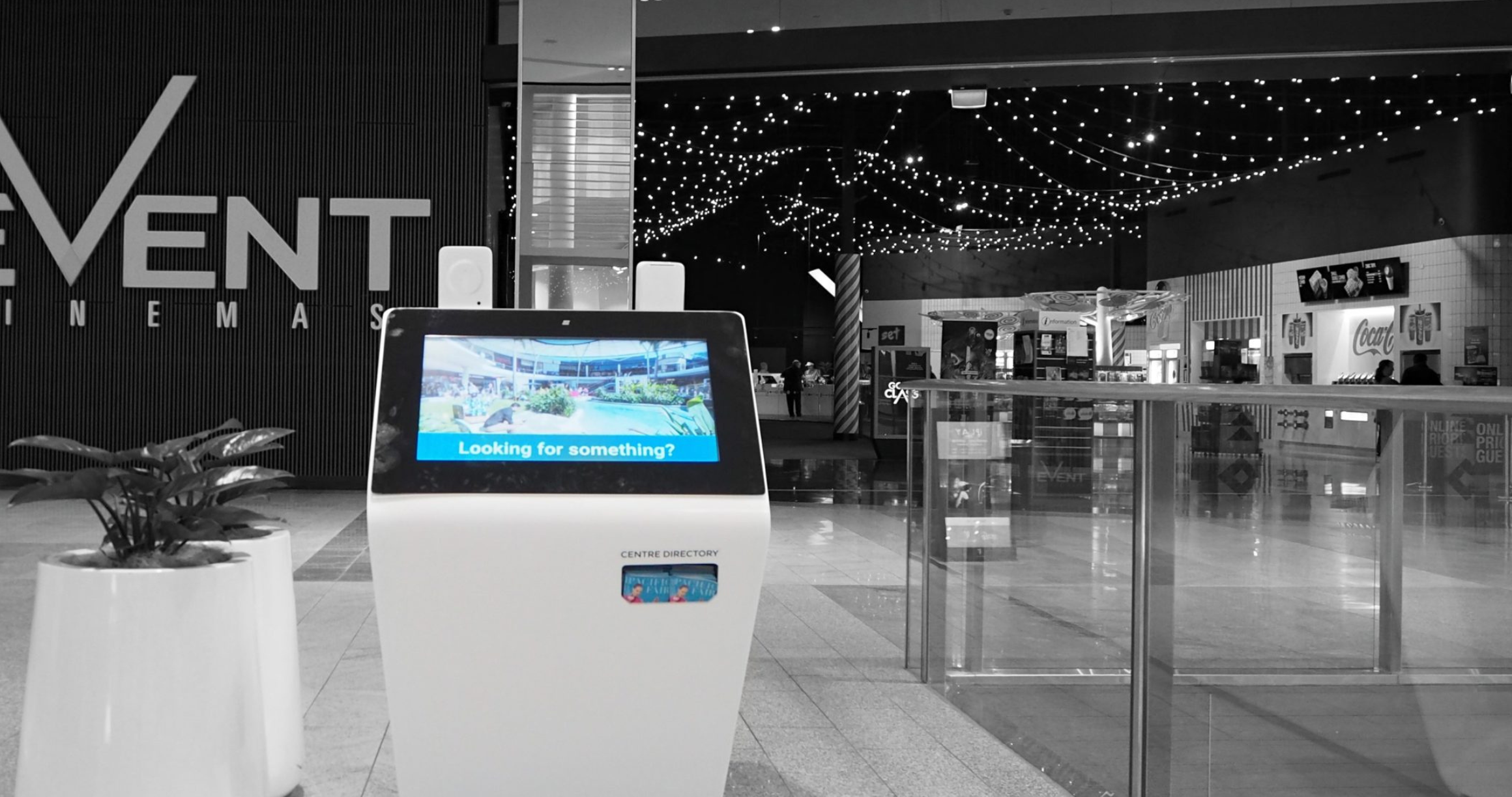 Touchscreen directory kiosk in a shopping mall or cinema lobby with the screen showing 'Looking for something?' and strings of lights hanging from the ceiling in the background.